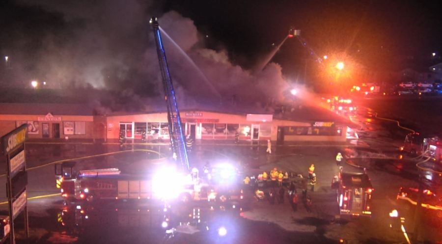 A ladder extends over a building. Firefighters spray water from above down at a commercial building with smoke billowing out of it.